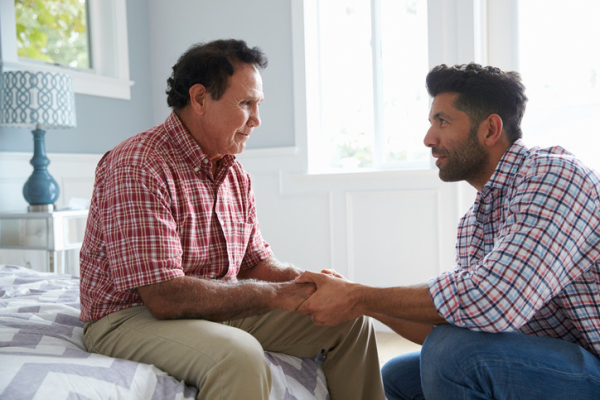Confused older father with dementia seated on bed, adult son kneeling, holding his hands and talking to him