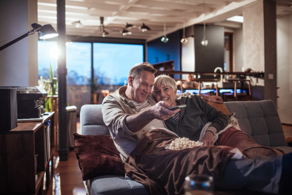 Smiling couple sitting on couch watching TV; man with short brown hair points remote, woman has white hair; bowl of popcorn rests on blanket 