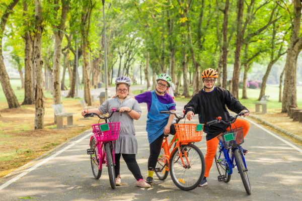 Three children with their bikes in a park with large green-leafed trees; the children, who are neurodiverse, are wearing helmets and grouped in a friendly way
